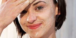 Esfoliante físico e químico. Foto mostra o rosto de uma mulher branca e cabelo preto com produto esfoliante branco sobre ele