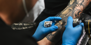 Dúvidas mais comuns tatuagens. Foto mostra um braço sendo tatuado. Tatuador está com luvas azuis e máscara