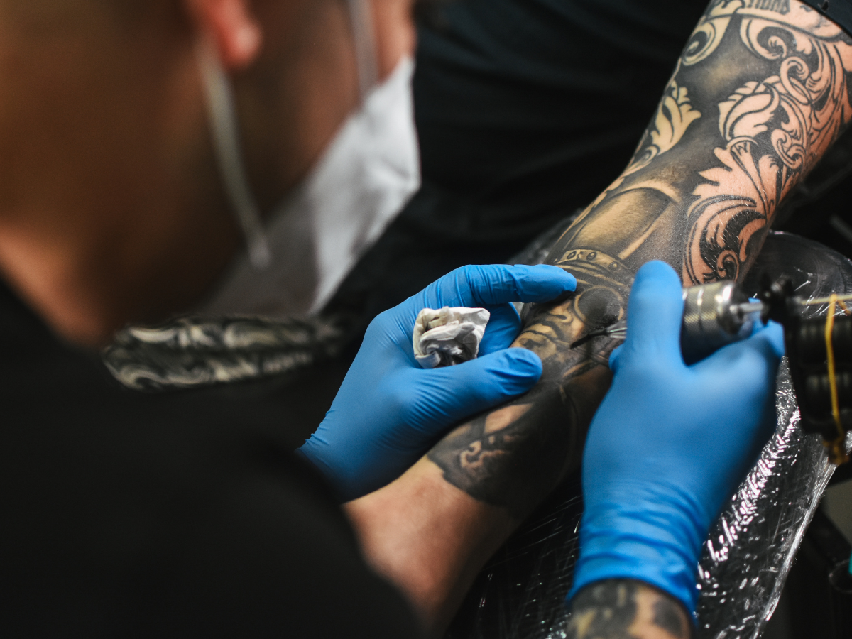 Dúvidas mais comuns tatuagens. Foto mostra um braço sendo tatuado. Tatuador está com luvas azuis e máscara