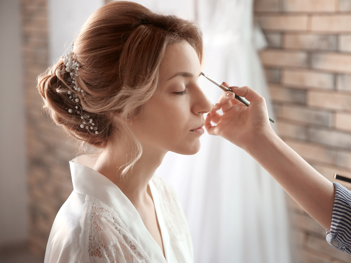 Penteados para noivas de cabelo longo. Foto mostra uma noiva loira com coque de acessório com pedraria. Ela está sendo maquiada.