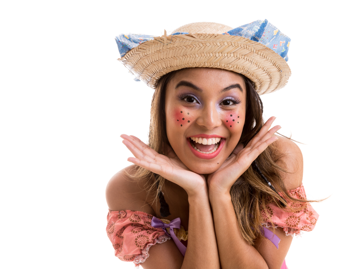 Maquiagem para festa junina. Foto mostra mulher sorrindo com sardinhas desenhadas na bochecha e chapéu junino