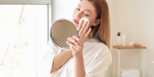Remover a maquiagem sem danificar a pele. Foto aparece mulher de blusa branca e cabelos castanhos retirando a maquiagem em frente ao espelho.