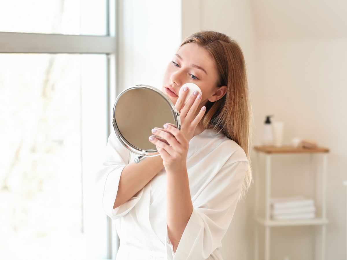 Remover a maquiagem sem danificar a pele. Foto aparece mulher de blusa branca e cabelos castanhos retirando a maquiagem em frente ao espelho.