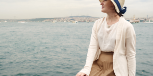 Saia midi no inverno. Foto mostra uma mulher com saia midi marrom, sentada sobre um estrutura em frente ao mar