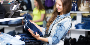Modelos de calças jeans; Foto mostra uma mulher branca, com cabelos lisos e jaqueta jeans com calça jeans nas mãos escolhendo em uma loja.