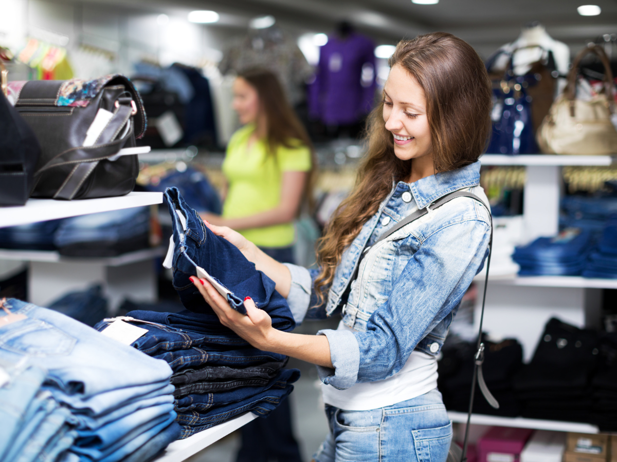 Modelos de calças jeans; Foto mostra uma mulher branca, com cabelos lisos e jaqueta jeans com calça jeans nas mãos escolhendo em uma loja.