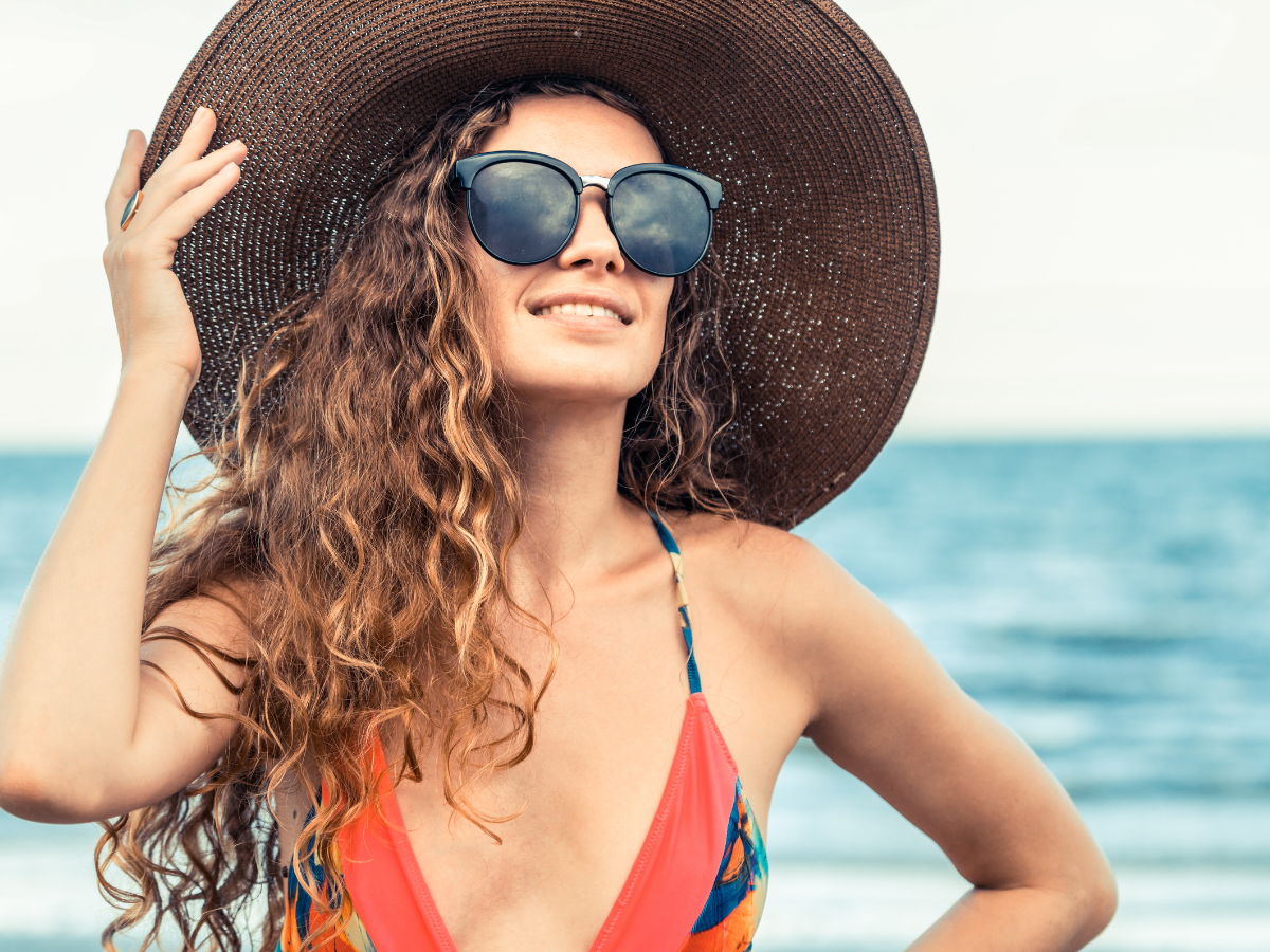 Cabelo no verão. Foto traz uma mulher sorrindo com cabelos cacheados e protegidos com um chapéu e óculos escuros