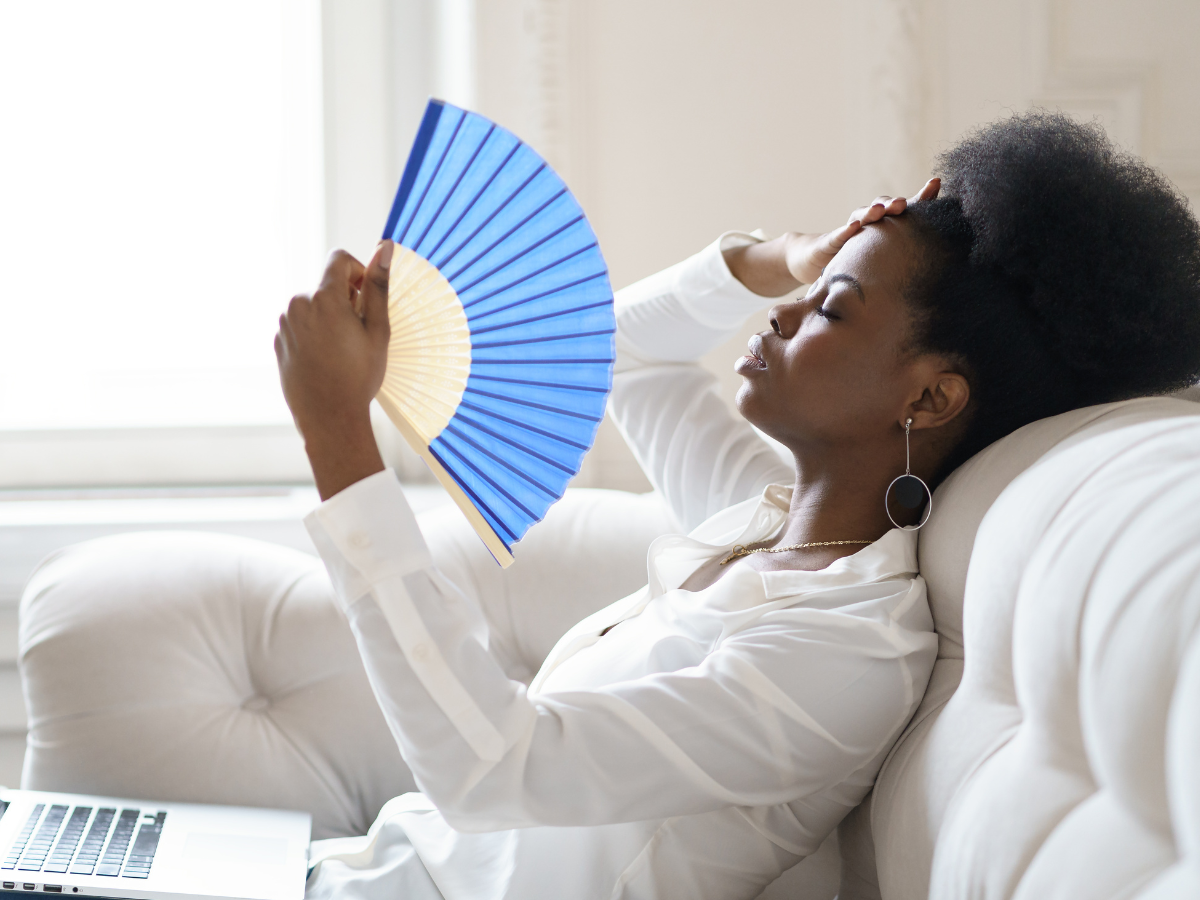 onda de calor. foto mostra mulher negra sentada sobre um sofá de cor branca se abanando com um leque de cor azul
